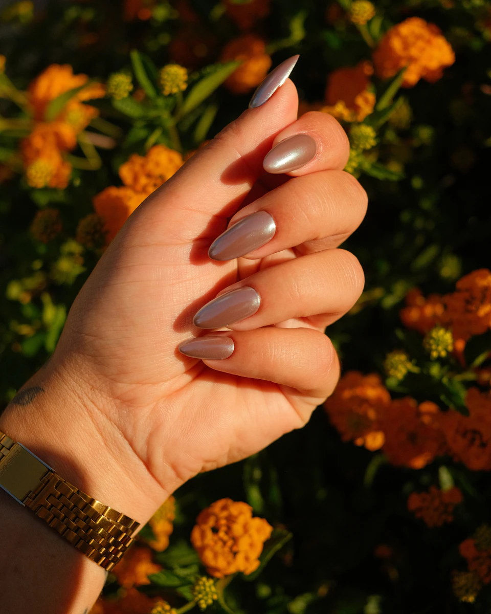 Uñas almendra en bronce cromado entre flores naranjas al atardecer